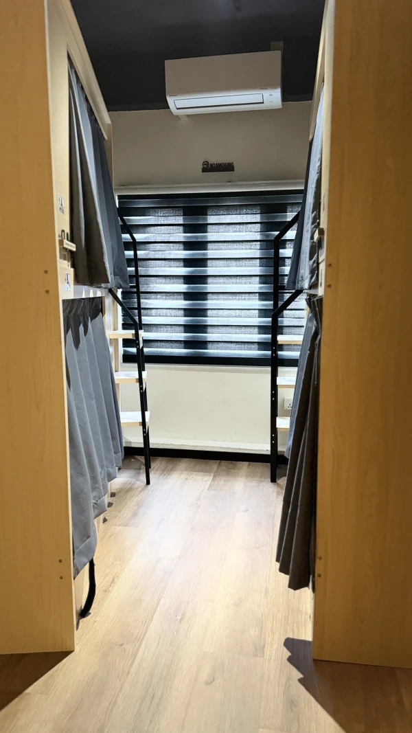 The aisle between two rows of pod bunks with grey curtains, leading to the window, in the female dormitory at Travelogue Guesthouse